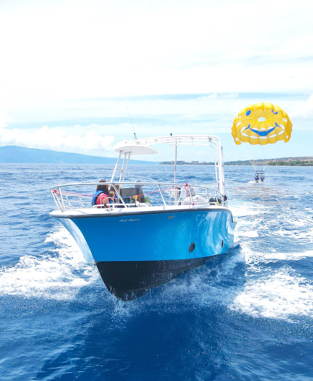 The blue parasail boat Chute Happens under way off Kaanapali, a pair of flyers dipping their feet in the water behind it