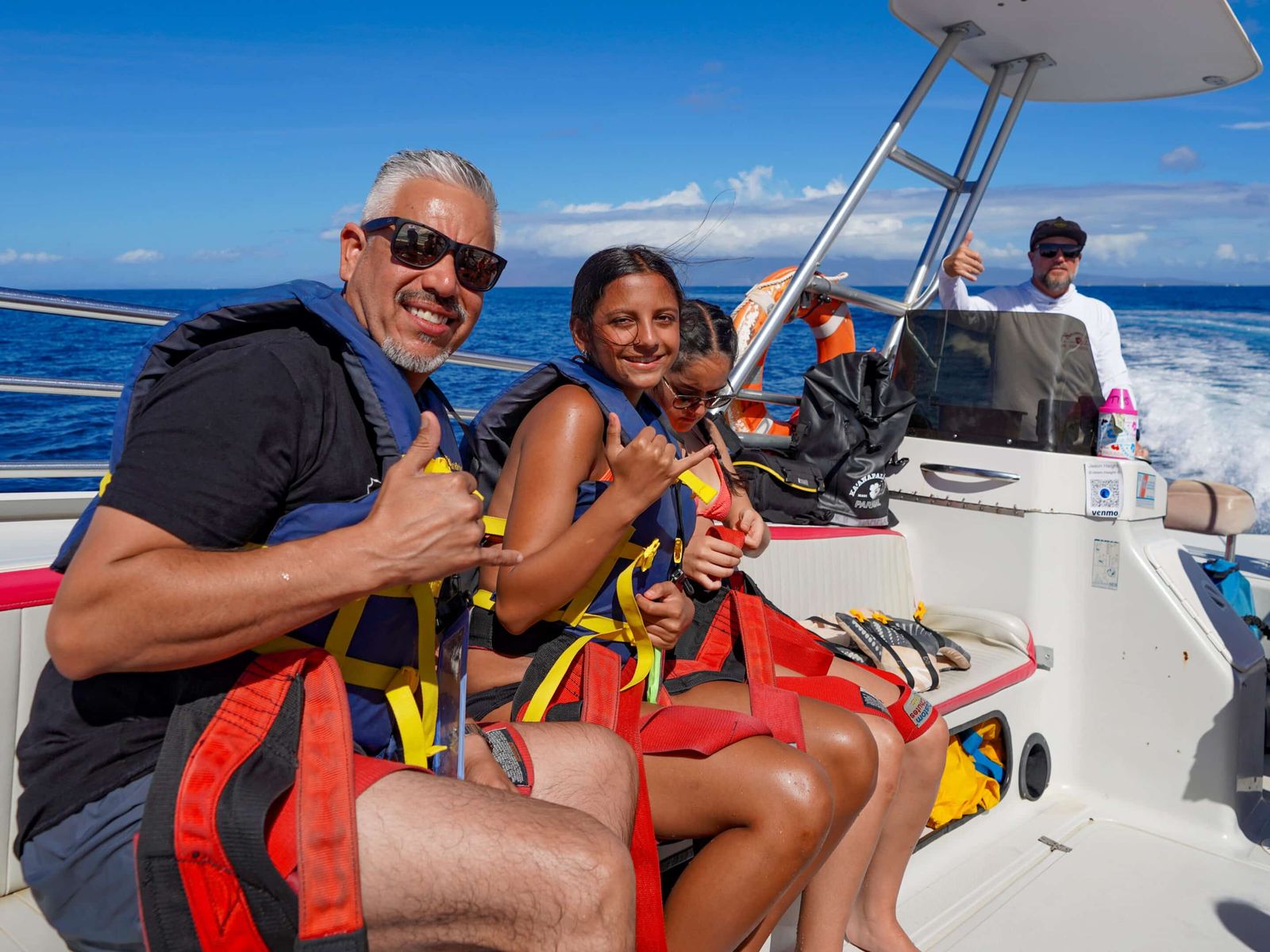 A father and two daughters in harnesses and life vests on the boat, throwing shakas, the captain at the helm behind them
