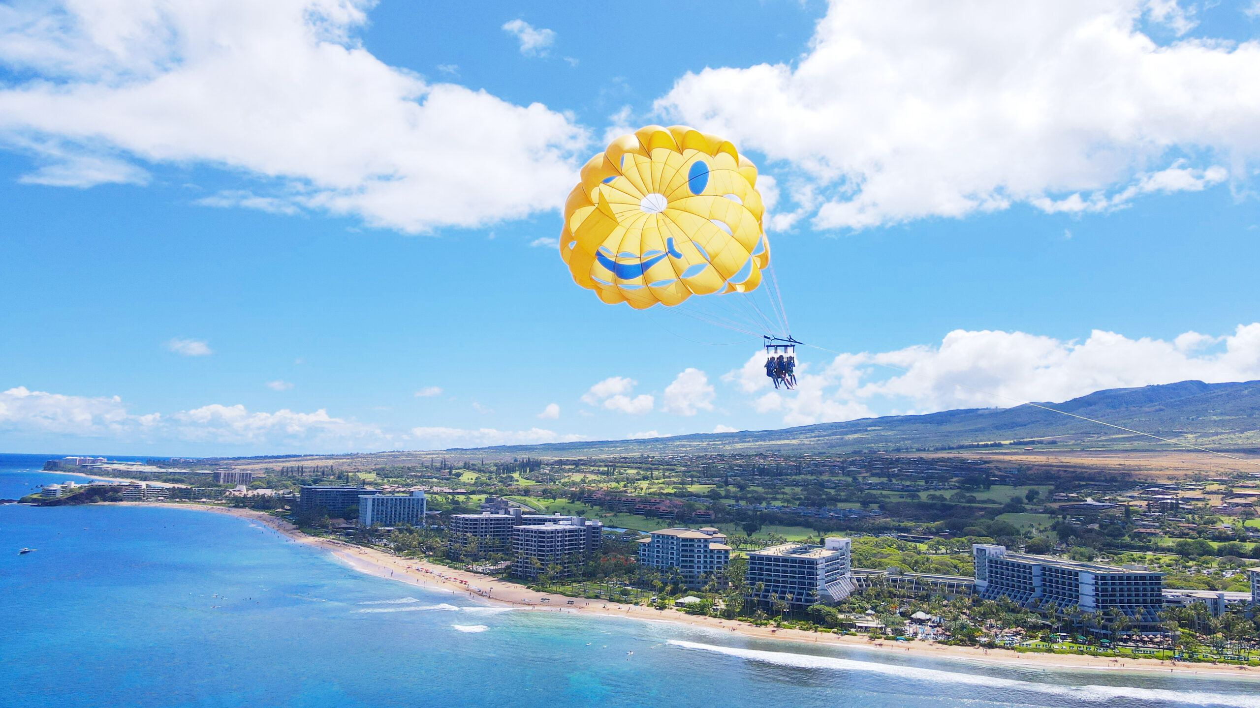 Three flyers under a yellow smiley-face parasail high above Kaanapali Beach, the resorts and the West Maui Mountains below